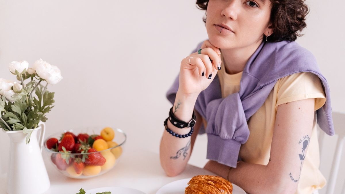 A stylish teenager with piercings enjoys a breakfast of croissant and fruit indoors.